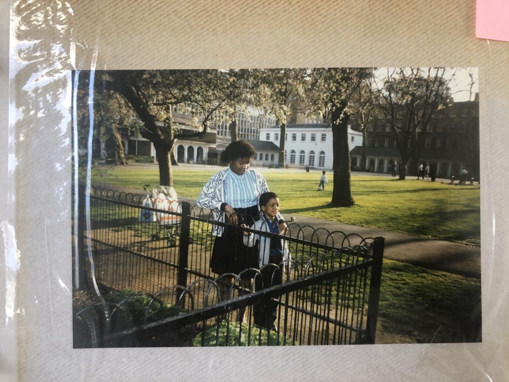 Photo of a photo in an album. A mother stands behind her son, holding a handrail in the middle of Coram's Fields park